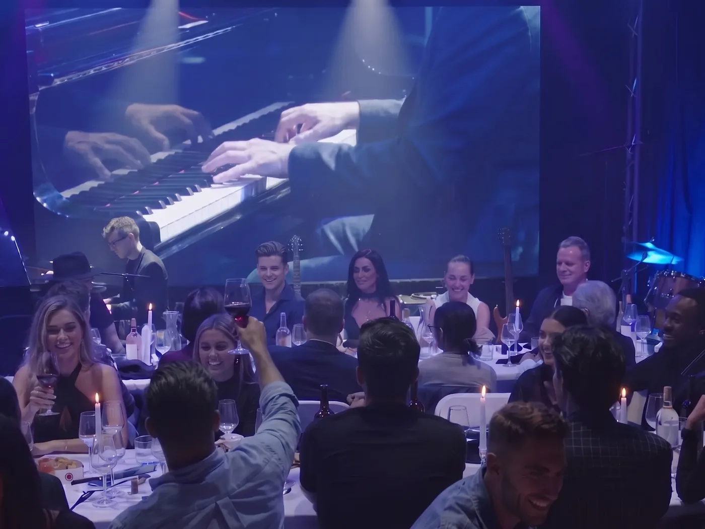 Guests at candlelit tables on the Live Room floor, the stage screen behind them showing a pianist’s hands