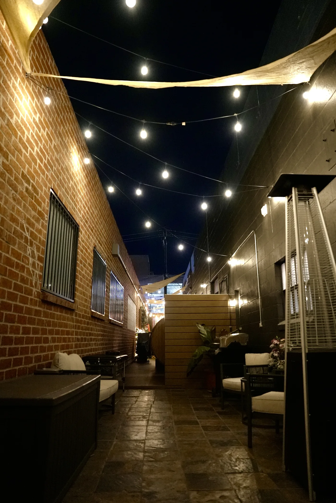 The patio at night, string lights strung between a brick wall and the neighbouring building, seating and a heater below
