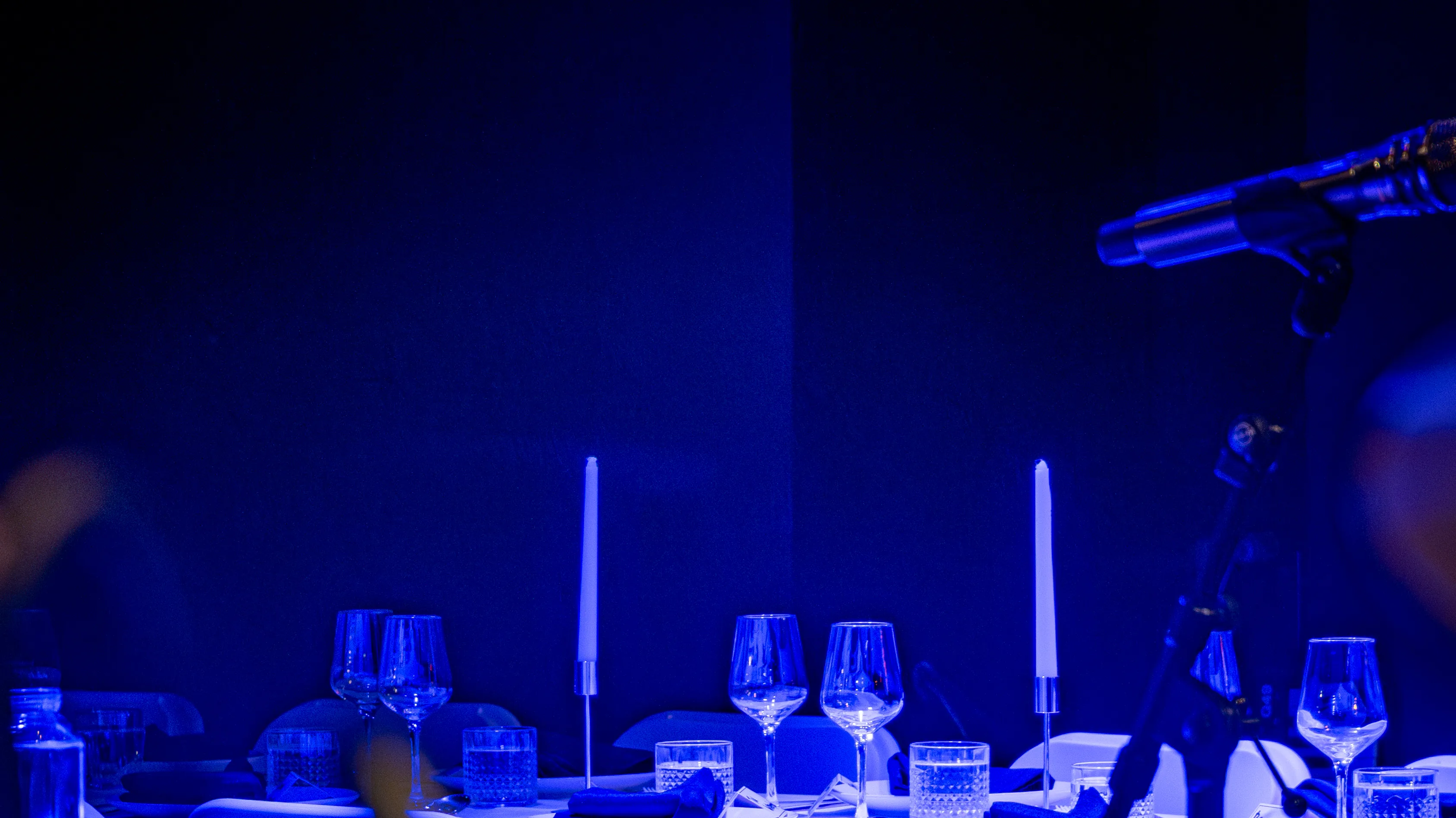 A long table on the Live Room floor set with tapered candles and cut crystal, lit deep blue, a microphone boom at the edge of frame