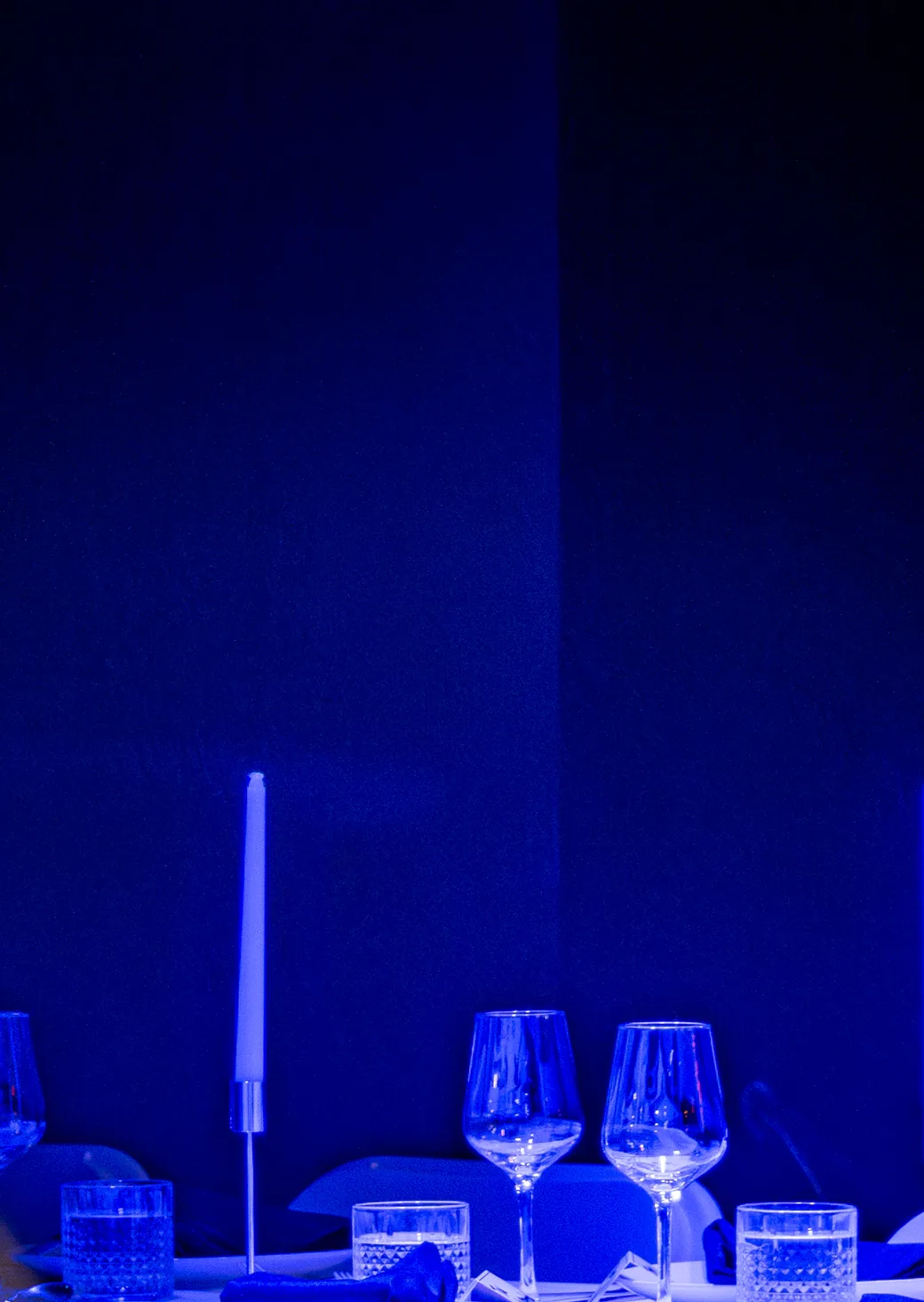 A dinner table under blue light on the Live Room floor: a tapered candle, wine glasses and cut-crystal tumblers