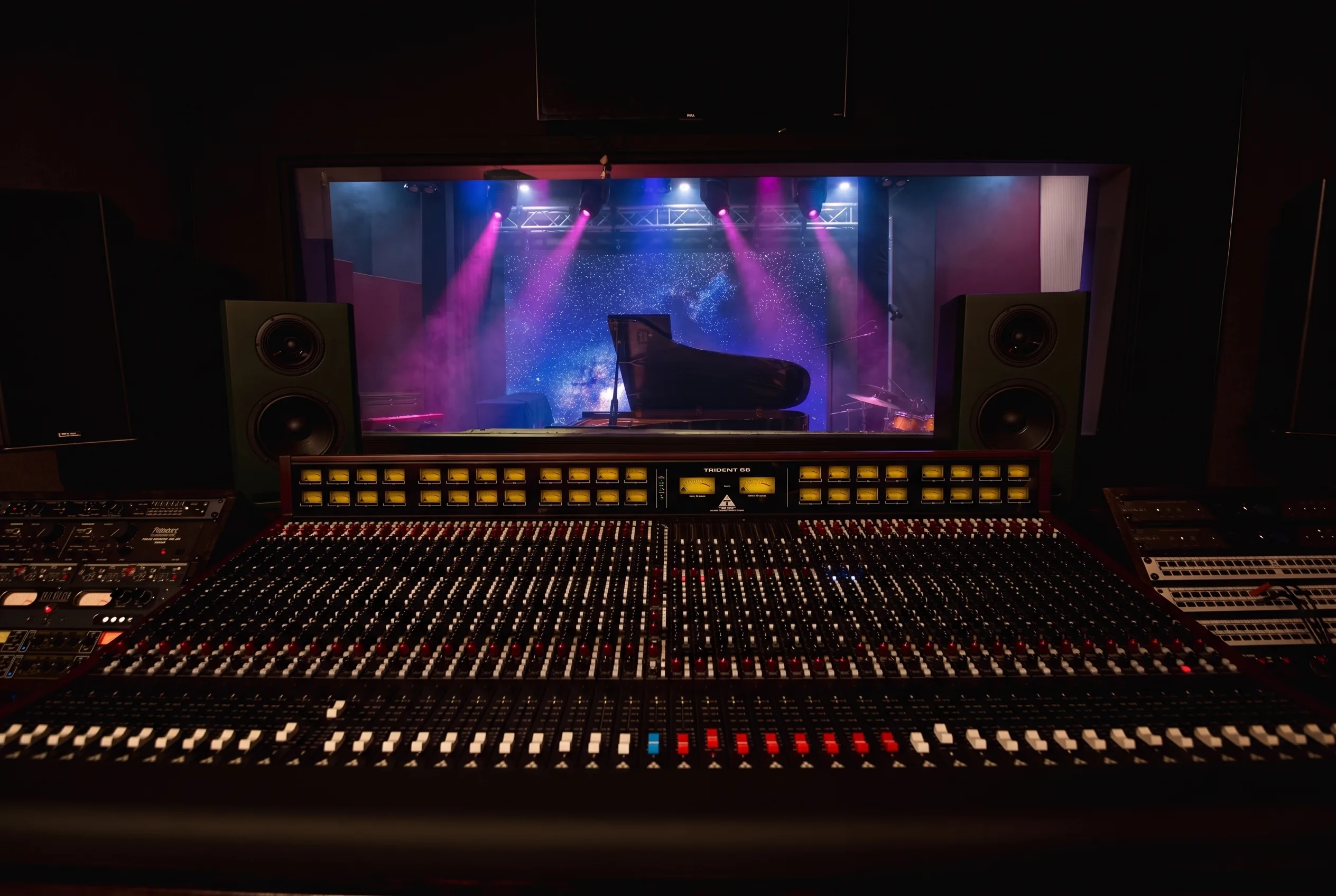 The Trident 88 console head-on in low light, the Live Room blazing beyond the control-room glass: starfield LED wall, magenta and blue beams, the grand piano on stage