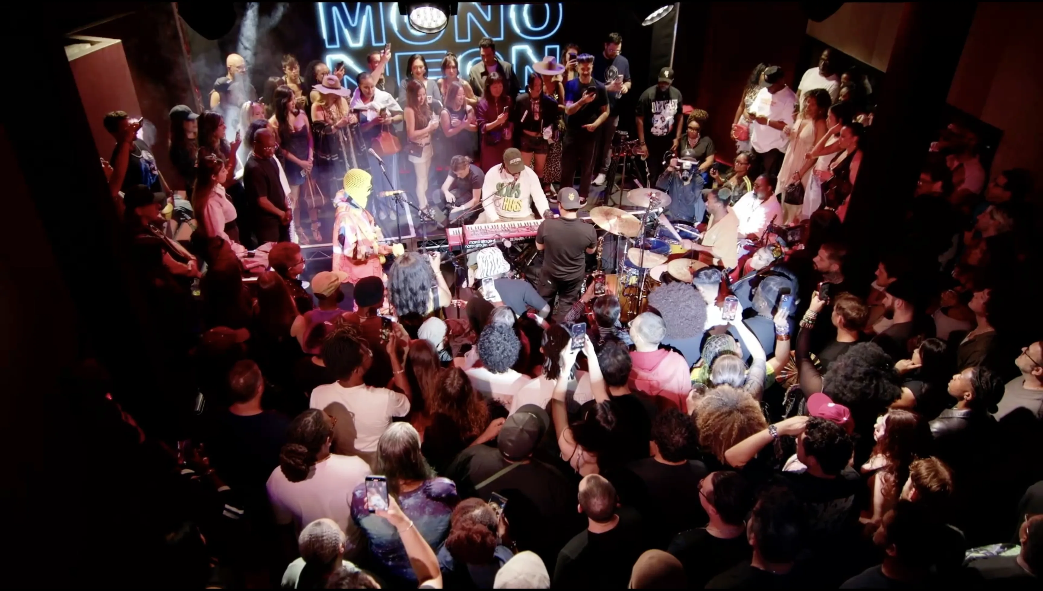 A packed room standing shoulder to shoulder around a band on the Live Room stage, phones up, haze in the lights