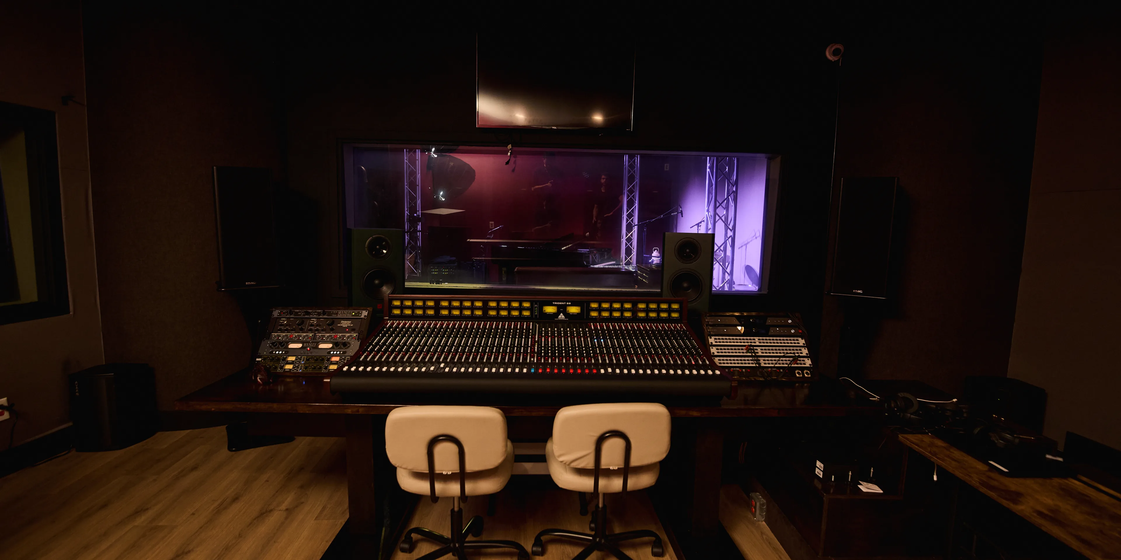 The Mixing Room facing the console, racks of outboard either side, the Live Room lit violet through the glass